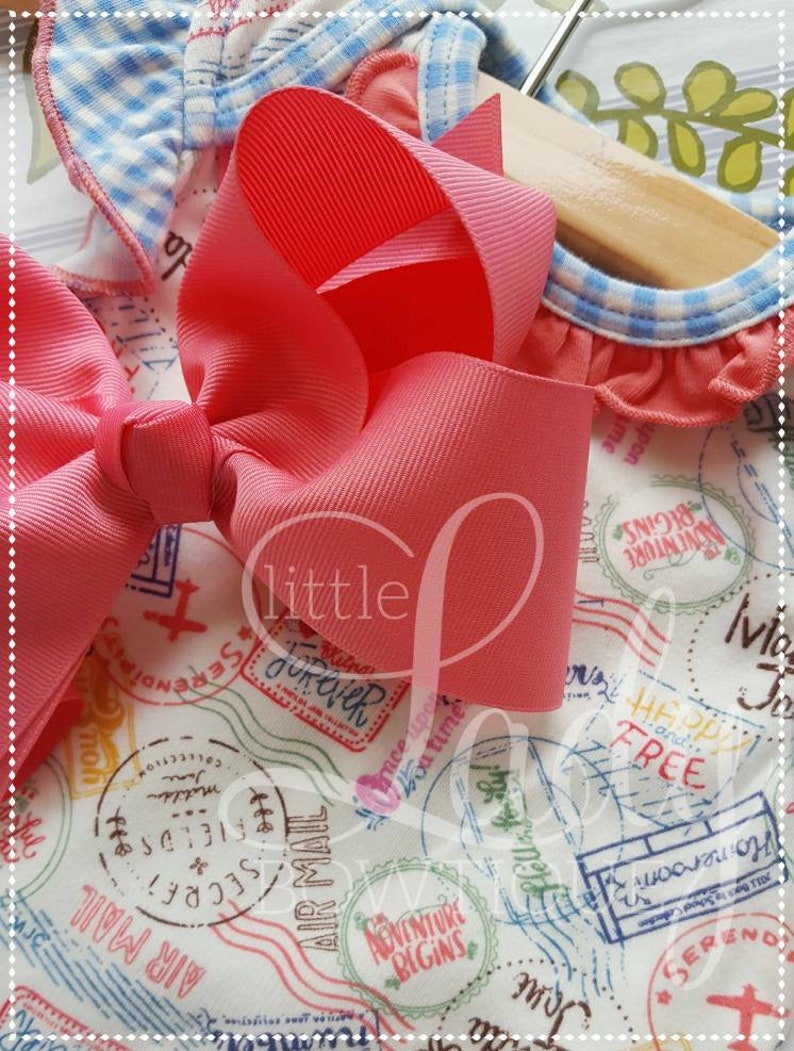 May include: A pink and white fabric with a blue and white gingham trim. The fabric has a pattern of stamps with words like "Adventure Begins", "Air Mail", and "Serendipity". A large pink bow is on top of the fabric.