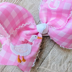 May include: A large pink gingham hair bow with white lace trim. The bow features an embroidered white goose with an orange beak and feet, wearing a pink bow. The bow has a white center knot and a monogrammed initial.