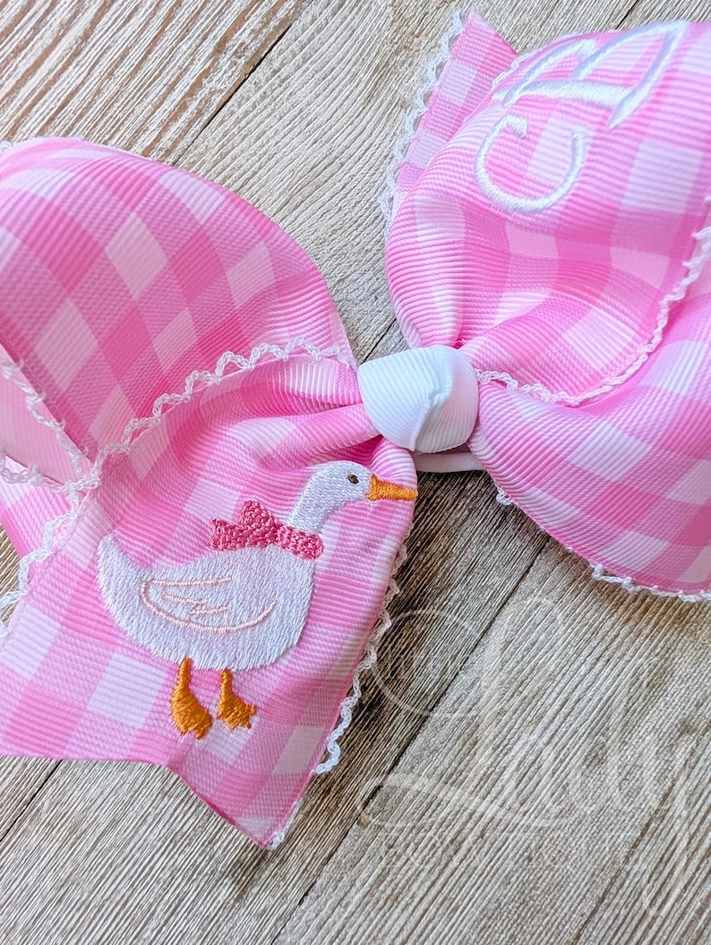 May include: A large pink gingham hair bow with white lace trim. The bow features an embroidered white goose with an orange beak and feet, wearing a pink bow. The bow has a white center knot and a monogrammed initial.