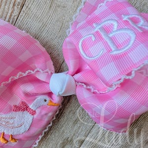 May include: A large pink and white gingham bow with white lace trim. The bow features an embroidered duck with a pink bow and the initials "CB" in white. The bow is tied with a white knot.