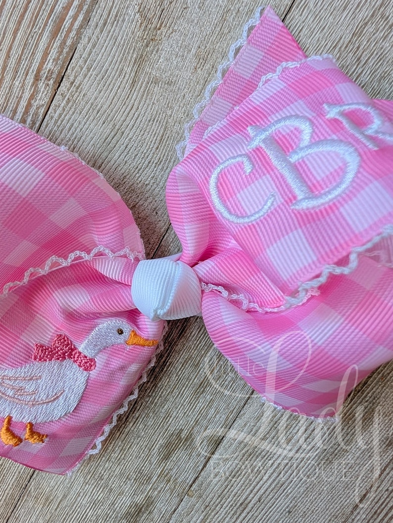 May include: A large pink and white gingham bow with white lace trim. The bow features an embroidered duck with a pink bow and the initials "CB" in white. The bow is tied with a white knot.