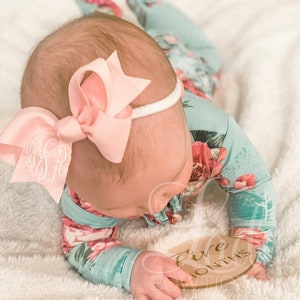 May include: A baby wearing a floral print onesie and a pink bow headband is holding a wooden sign that says "Five Months".