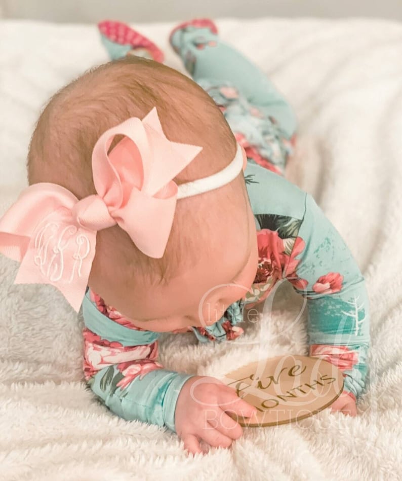 May include: A baby wearing a floral print onesie and a pink bow headband is holding a wooden sign that says "Five Months".