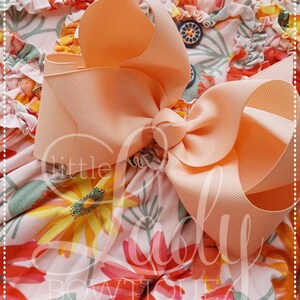 May include: A peach-colored bow with a large, full knot, tied on a pink and white floral fabric with orange, yellow, and green accents.
