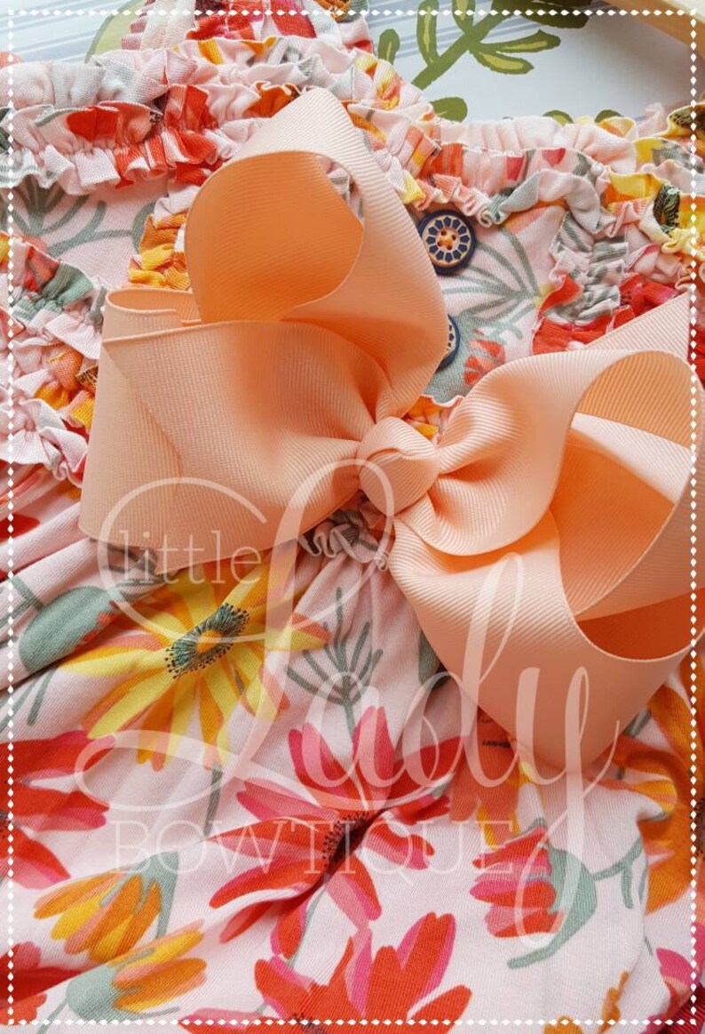 May include: A peach-colored bow with a large, full knot, tied on a pink and white floral fabric with orange, yellow, and green accents.