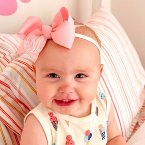 May include: A baby girl wearing a pink bow headband with a white monogram on a white background. The headband is on top of her head and the bow is centered. She is wearing a white sleeveless shirt with a colorful ice cream cone pattern. She is smiling and looking at the camera.