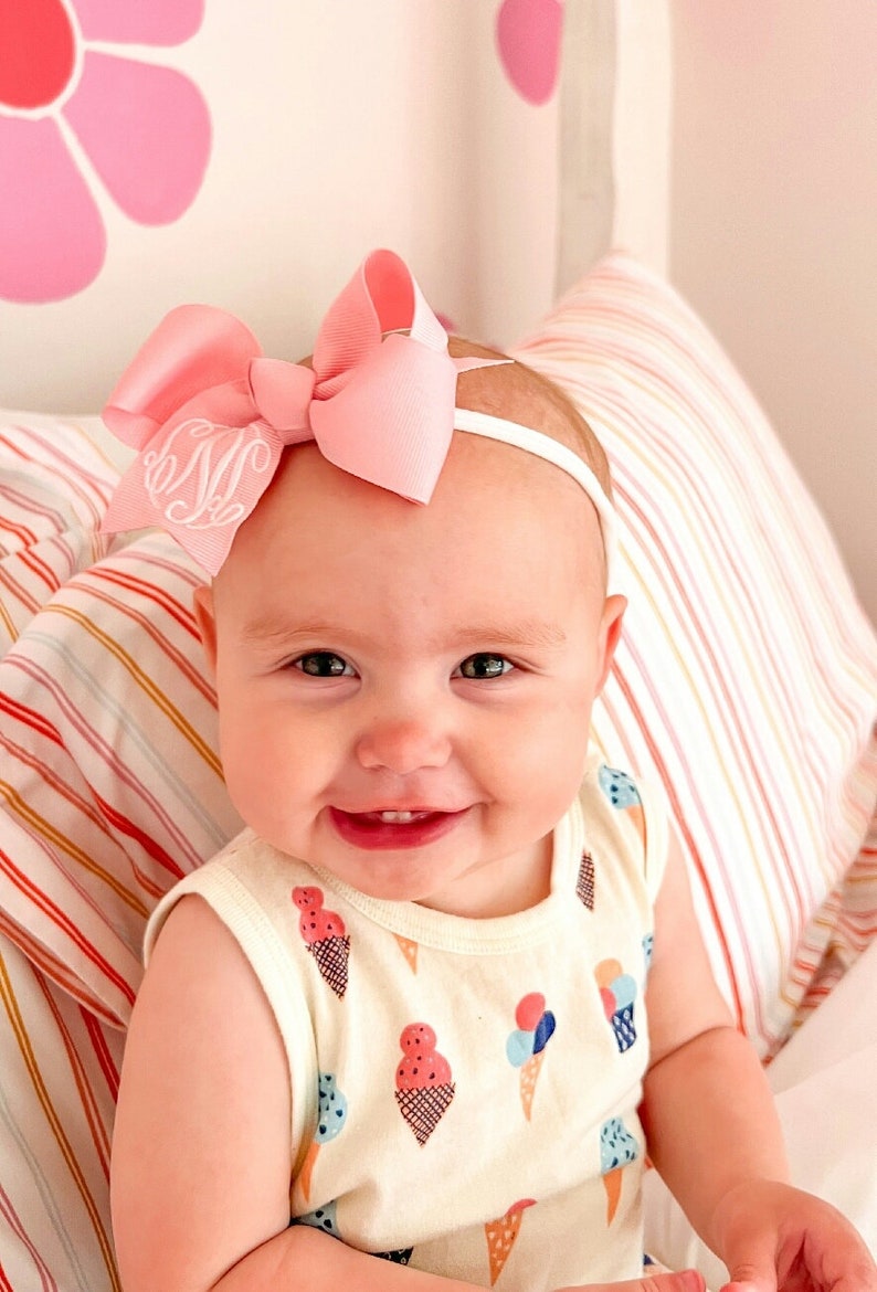 May include: A baby girl wearing a pink bow headband with a white monogram on a white background. The headband is on top of her head and the bow is centered. She is wearing a white sleeveless shirt with a colorful ice cream cone pattern. She is smiling and looking at the camera.
