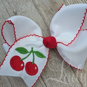 Può includere: Un fiocco per capelli in nastro gros-grain bianco con bordo cucito rosso e un nodo centrale rosso. Il lato sinistro del fiocco presenta un ricamo di due ciliegie rosse con gambi e foglie verdi su uno sfondo bianco.