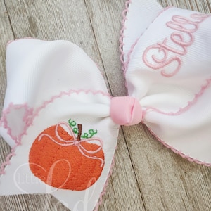 Könnte beinhalten: Ein weißer Haarreifen mit rosa Besatz und einem gestickten Kürbisdesign. Die Schleife ist mit dem Namen "Stella" in rosa Garn personalisiert.