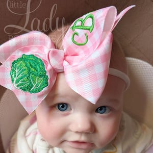 May include: A pink and white gingham bow with a green embroidered cabbage and green embroidered initials. The bow is attached to a white headband. The image also includes the text "little Lady Boutique".