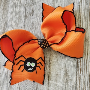 Könnte beinhalten: Orange-schwarzes Halloween-Haarband mit einer schwarzen Spinne mit grünen Augen, die auf einer Seite gestickt ist. Die Schleife hat einen schwarzen gewellten Rand.