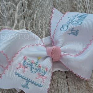 May include: A white grosgrain ribbon hair bow featuring pink scalloped stitched edges and a pink centre knot. The bow is embroidered with a light blue birthday cake with pink roses and the light blue text "Ava's 1st Birthday".