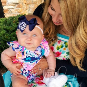 May include: A baby girl wearing a white headband with a navy blue bow and a white monogram, a floral print onesie, and white booties with a unicorn design. The baby is being held by a woman wearing a floral print dress.