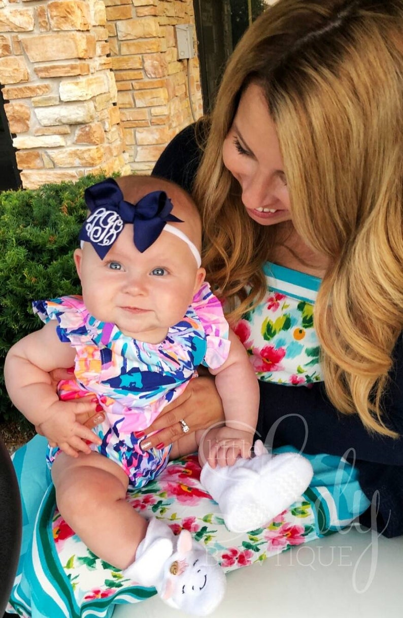 May include: A baby girl wearing a white headband with a navy blue bow and a white monogram, a floral print onesie, and white booties with a unicorn design. The baby is being held by a woman wearing a floral print dress.