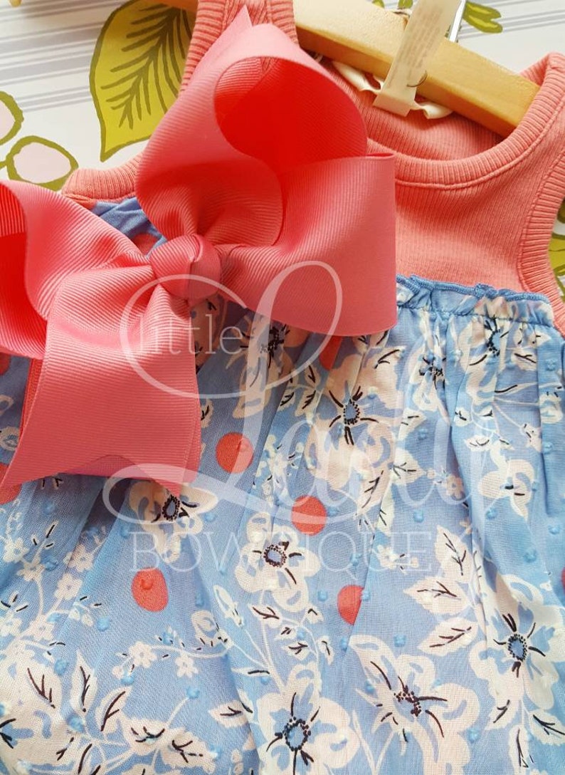 May include: A light blue and coral sleeveless dress with a white floral print and coral polka dots. A large coral bow is tied at the top of the dress.