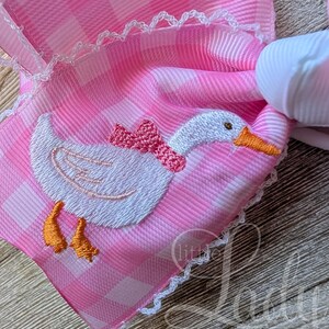 May include: Pink gingham hair bow with a white embroidered goose wearing a pink bow. The bow has white lace trim and a white ribbon base. The goose has an orange beak and feet. The bow is a decorative hair accessory.