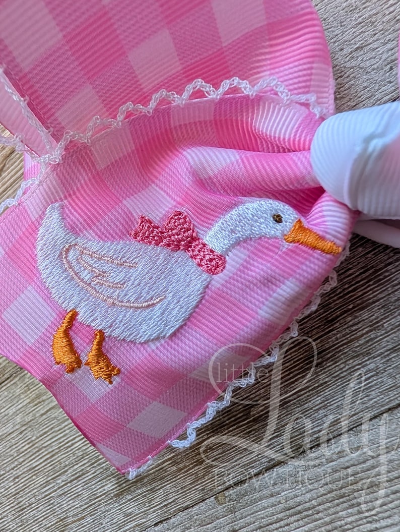 May include: Pink gingham hair bow with a white embroidered goose wearing a pink bow. The bow has white lace trim and a white ribbon base. The goose has an orange beak and feet. The bow is a decorative hair accessory.