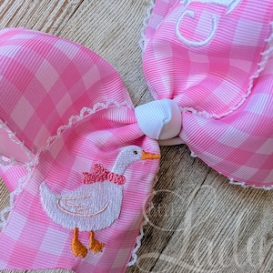 May include: A large pink gingham hair bow with white lace trim. The bow features an embroidered white duck with an orange beak and feet, and a pink bow. The bow is tied with a white knot and has a white embroidered initial.