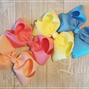 May include: Four large hair bows in shades of orange, yellow, and blue. The bows are made of grosgrain ribbon and are arranged in a row on a wooden surface.