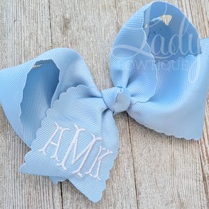 May include: A light blue hair bow made of ribbed fabric with scalloped edges. The bow features white embroidered monogram letters HMK on one of the tails. The texture of the ribbon is visible throughout the surface of the light blue bow.