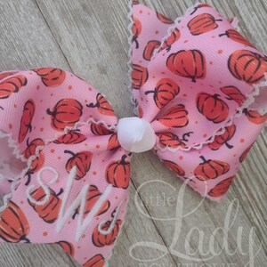 May include: A pink hair bow with a white lace trim and a pumpkin print. The bow has the initials "SW" embroidered in white on the front.