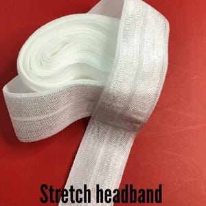 May include: White stretch headband, partially rolled, against a red background. The headband is made of a textured, elastic material. The text "Stretch headband" is printed in black at the bottom of the image.