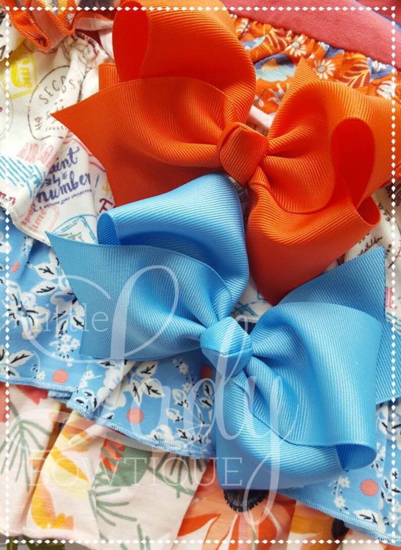 May include: Two hair bows, one orange and one light blue, are shown on a patterned fabric background. The bows are made of a grosgrain ribbon.