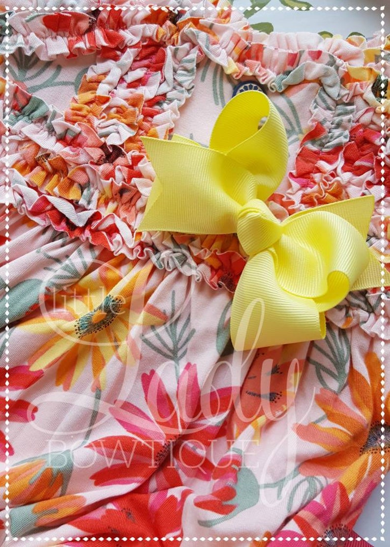May include: A pink and white floral print dress with a yellow bow. The dress has a ruffled neckline and a floral pattern with orange, pink, and yellow flowers.