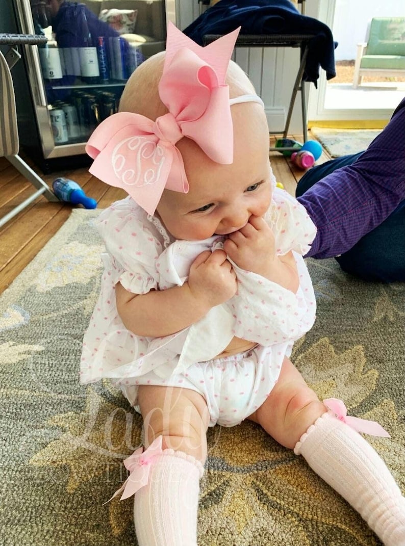 May include: A baby girl wearing a white dress with pink polka dots, a pink bow headband with a white embroidered letter 'S', and pink knee-high socks with a pink bow.
