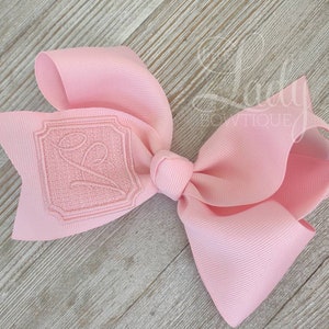 Puede incluir: Un lazo para el pelo rosa con una inicial bordada en un marco cuadrado. La inicial es una "E" en cursiva de color rosa claro.