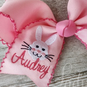 以下が含まれることがあります： ピンクのヘアボウに、白い刺繍のウサギの顔とピンクの糸で「Audrey」という名前が刺繍されています。