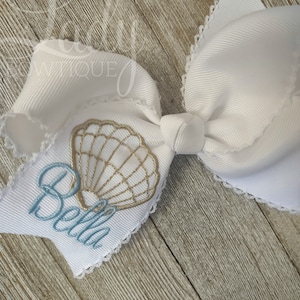 May include: White bow with a lace trim and a gold seashell with the name Bella embroidered in blue thread.