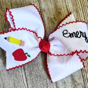 May include: A white hair bow with a red scalloped edge and a red centre knot. The left side features an embroidered yellow pencil and a red apple. The right side features the name Emery embroidered in black script lettering on the white fabric.