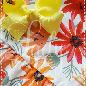 May include: A yellow bow with a white background featuring a floral pattern with orange, red, and green flowers. The text "little lady boutique" is visible in the image.