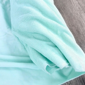 May include: A soft, mint green fabric with a plush, velvety texture. The material is folded over itself, showcasing the dense, short pile surface and the raw, unfinished edges of the textile. The light color provides a consistent tone.