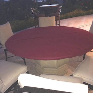 Puede incluir: Un mantel redondo de color burdeos cubre una mesa redonda grande de piedra con base de pedestal. Cuatro sillas con cojines blancos rodean la mesa. Las sillas tienen marcos de metal negro.