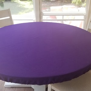 Puede incluir: Un mantel redondo de color morado drapeado sobre una mesa blanca con cuatro sillas blancas. El mantel es liso y sin arrugas.