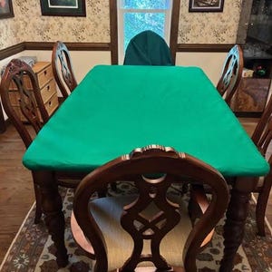 May include: A rectangular dining table covered with a solid bright green fabric cloth, surrounded by four dark brown wooden chairs with intricate carved backrests and light-colored upholstered seats, positioned on a patterned area rug.