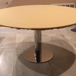 Puede incluir: Un mantel de mesa redondo de color beige cubre una mesa redonda de cromo con una base de pedestal simple. La mesa est&aacute; en una habitaci&oacute;n con un suelo de baldosas beige claro.
