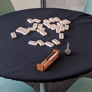 Puede incluir: Una mesa negra cubierta con un mantel negro con un juego de fichas de domin&oacute; blancas esparcidas sobre la superficie. Un peque&ntilde;o &aacute;baco de madera con cuentas marrones est&aacute; sobre la mesa, junto con un llavero plateado.
