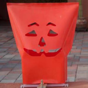 May include: A bright orange Halloween decoration shaped like a jack-o'-lantern. The decoration has cut-out eyes, nose, and a smiling mouth. The material appears to be a lightweight fabric or plastic, suitable for outdoor display during the fall season.