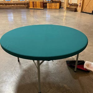 May include: A round, green table cloth covers a folding table with silver legs. The table is in a large, empty room with wooden walls and a concrete floor.