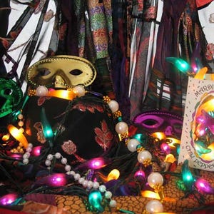May include: A festive display featuring Mardi Gras masks, strings of pearls, and colorful chili pepper lights. A box of lights is visible, with the words "Mardi Gras Chili Pepper Lights" printed on it. The scene is set against a backdrop of patterned fabrics.