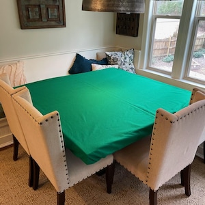 May include: A green tablecloth covers a square table with four beige chairs. The chairs have dark brown legs and are upholstered in a beige fabric with dark brown nailhead trim. The table is set in a dining room with a window and a light fixture hanging above.