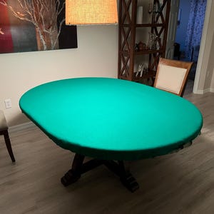 May include: Oval-shaped table with a dark wooden base and a vibrant green felt top. The table is set in a room with hardwood floors and a neutral color palette. The table's dimensions are approximately 60 inches in length and 40 inches in width.