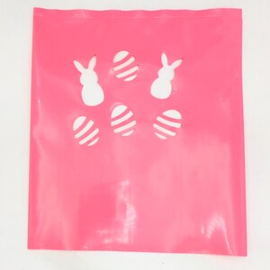 May include: A bright pink Easter-themed gift bag with cut-out designs of white Easter eggs and bunny silhouettes. The bag is made of a shiny, smooth material and is perfect for Easter gifts.