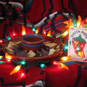 May include: A red and green chili pepper string of lights with a ceramic bowl and chips. The lights are shaped like chili peppers and are lit up. The bowl is filled with tortilla chips. The package for the lights says "Fiesta Hots Chili Pepper Lights".