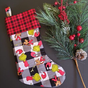 May include: A Christmas stocking with a black and white checkered pattern and a red and black buffalo plaid cuff. The stocking features a repeated pattern of the Grinch and Max the dog.