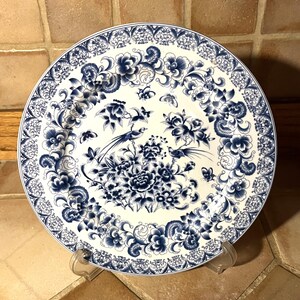 May include: A round ceramic plate featuring a white base with intricate blue floral patterns, scrolling foliage, and butterflies. The central design depicts two long-tailed birds perched among blooming flowers, surrounded by a decorative border.