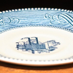 May include: A blue and white ceramic plate with a decorative border and a central image of a steamboat. The plate is perfect for serving desserts or snacks.